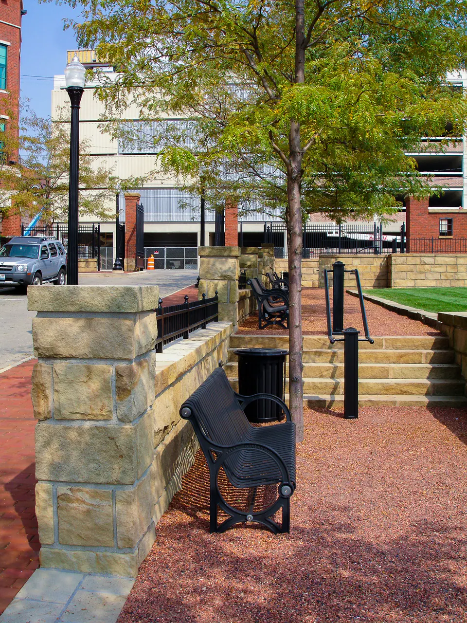 Stone pier, metal railing, bench, brick paving, lawn, trees, and commercial streetscape lighting.